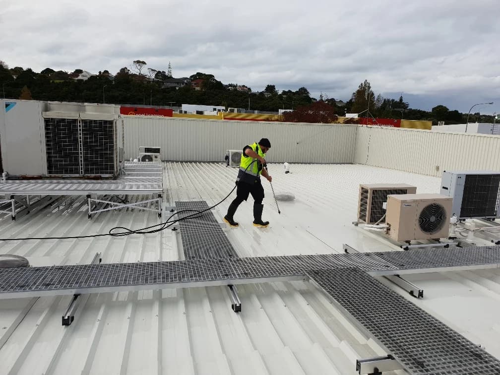 Technician washing a white commercial metal roof with a spray wand, working between rooftop HVAC units