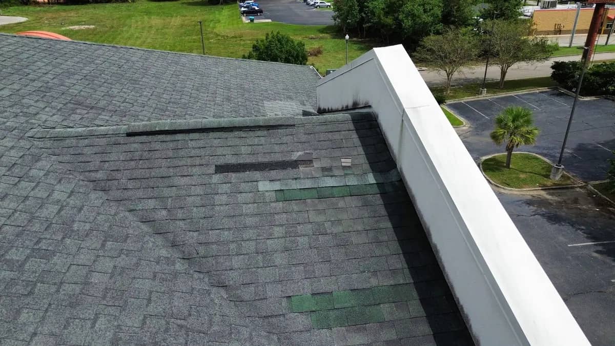 Close aerial view of displaced and missing shingles along a hip beside a parapet wall