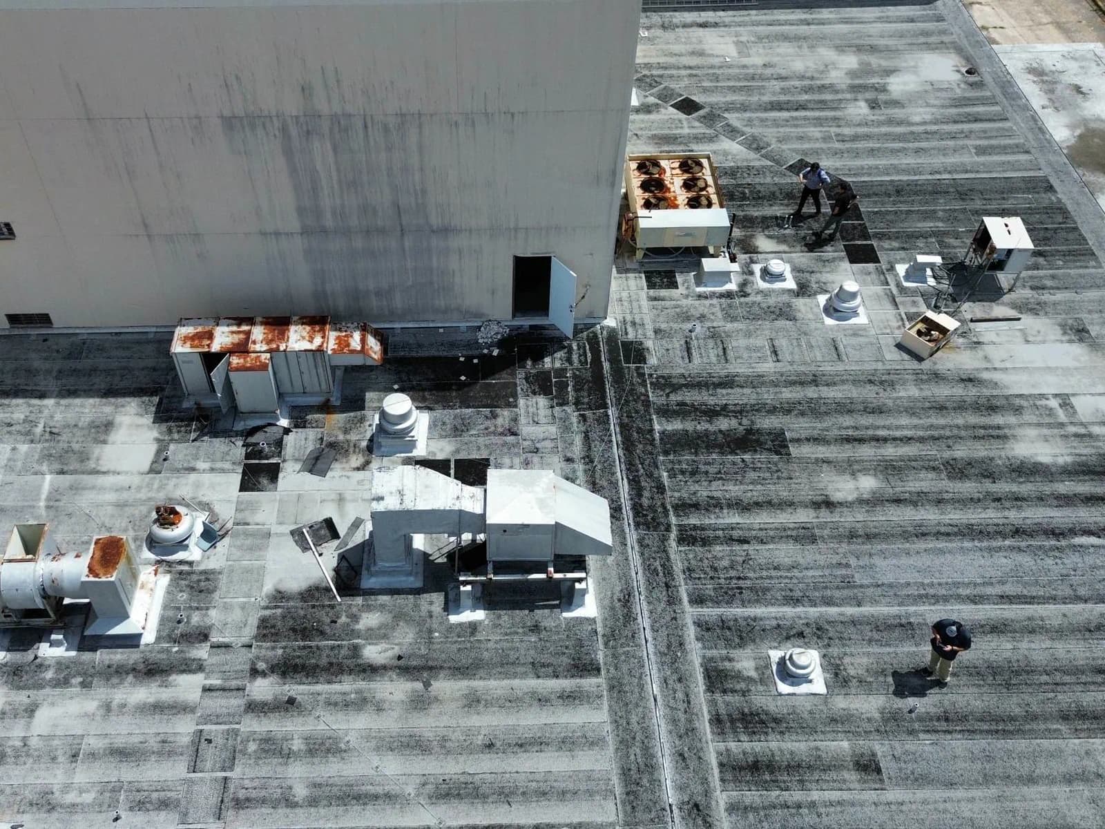 Aerial view of a Southeast Roofing crew walking an aged commercial flat roof during a maintenance survey, past rusted rooftop units and earlier patch repairs